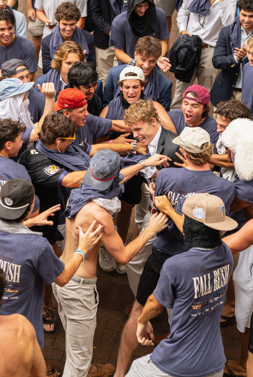 A group of young people in "Fall Rush" shirts and casual attire are gathered outdoors, laughing and embracing each other in a lively, energetic scene.
