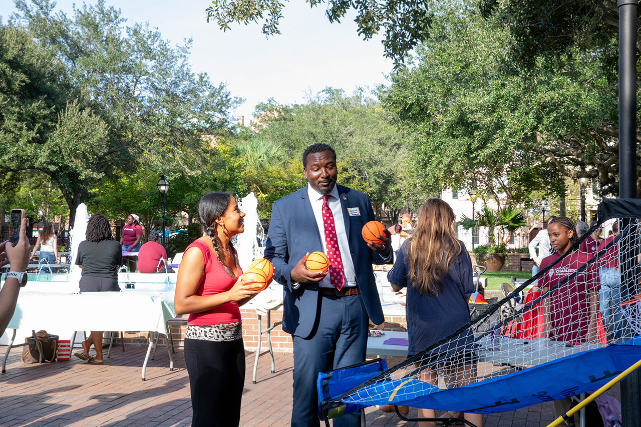A group of people, including a person in a suit and two others holding basketballs, stand outdoors near a trampoline net in a park setting.