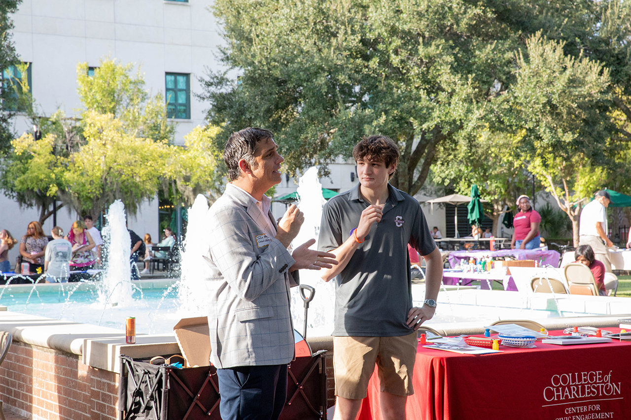 Two individuals engage in conversation outdoors near a fountain, with a red table bearing “College of Charleston” signage and event attendees visible in the background.