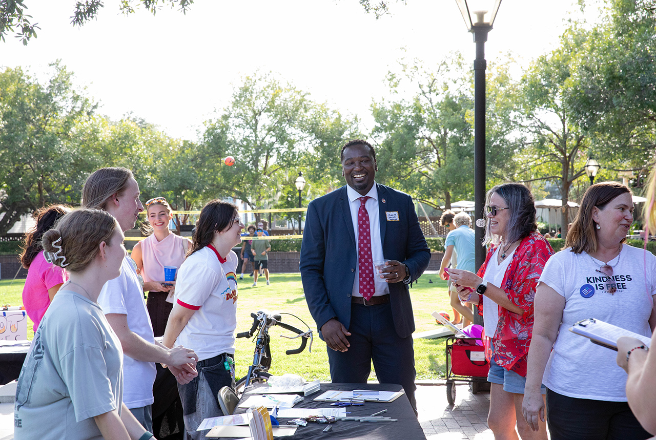 A group of people gather outdoors at a sunny event, engaging in conversation around a table with materials and a bicycle nearby.