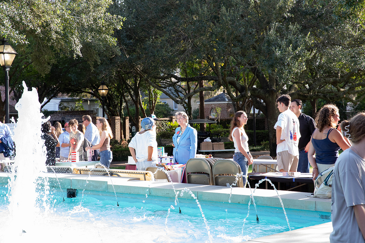 A group of people gather around a fountain in a park-like setting, with trees and tables in the background.