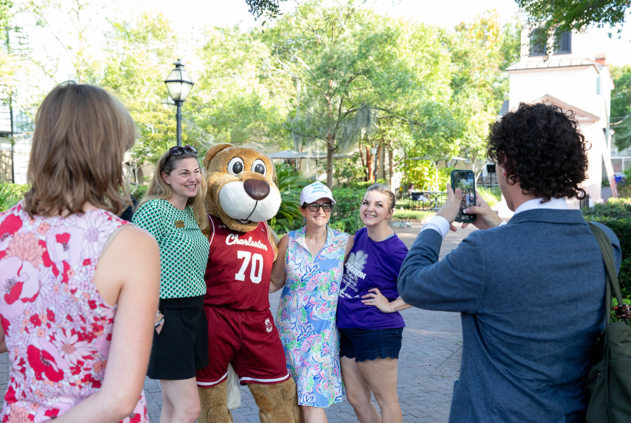 A group of people, including a person in a large bear mascot costume, pose for a photo taken by another individual holding a smartphone outdoors.