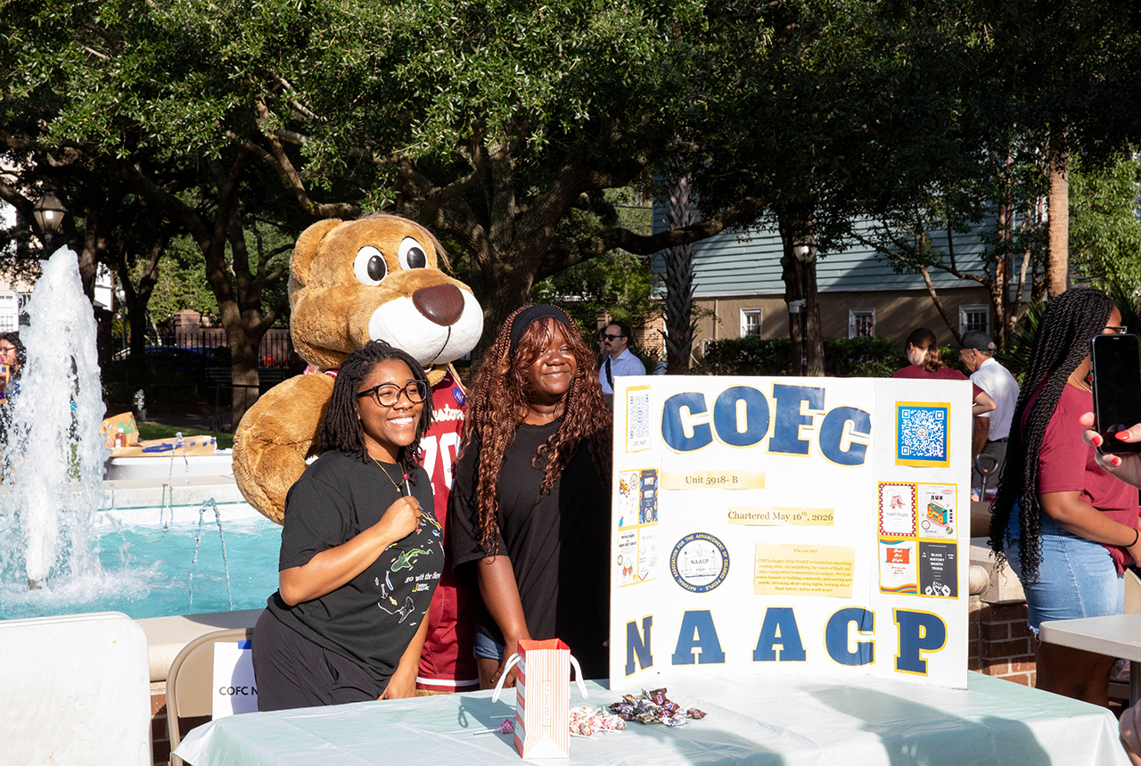 Two individuals smile beside a large teddy bear at an outdoor event, standing next to a table with a sign for COFC NAACP.