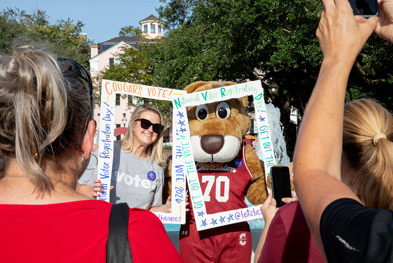 A person in a cougar mascot costume stands with a smiling individual inside a handmade frame that reads “Cougars Vote!” and “National Voter Registration Day!” while others take photos.