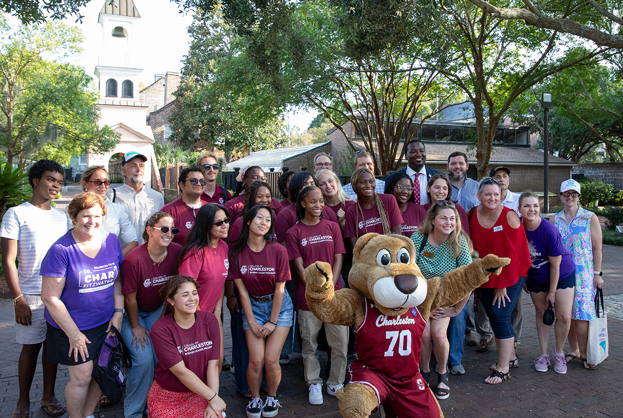 A group of people, including a mascot, gather outdoors in front of a historic church, many wearing maroon shirts with "Charleston" branding.