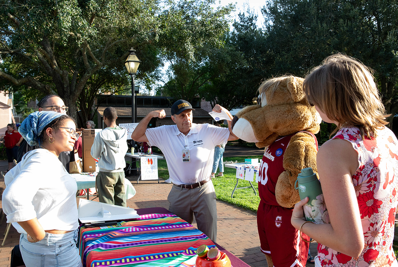 A person in a mascot costume interacts with others at an outdoor event, while another individual flexes their arms near a colorful table.