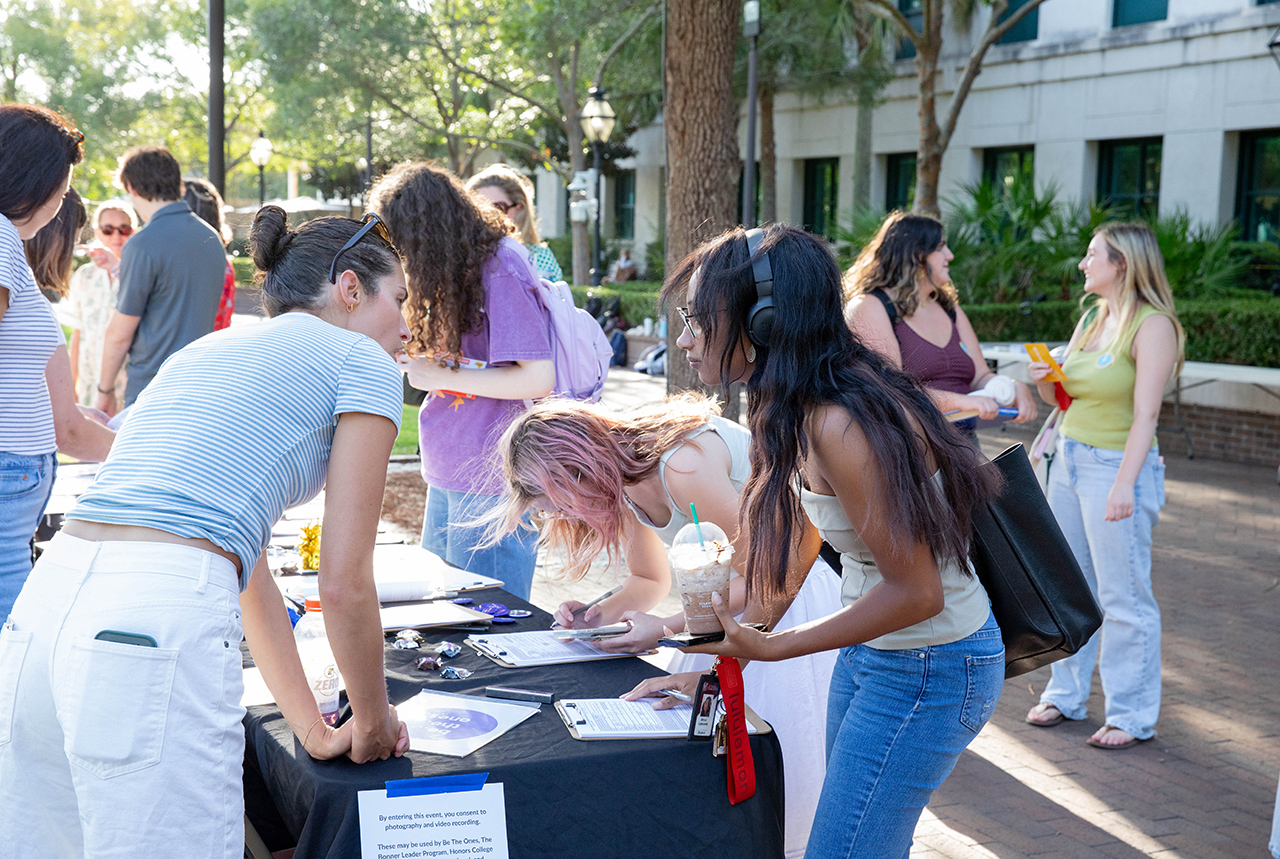 Several individuals gather around a table outdoors, engaging with materials and interacting with each other in a sunny, tree-lined setting.