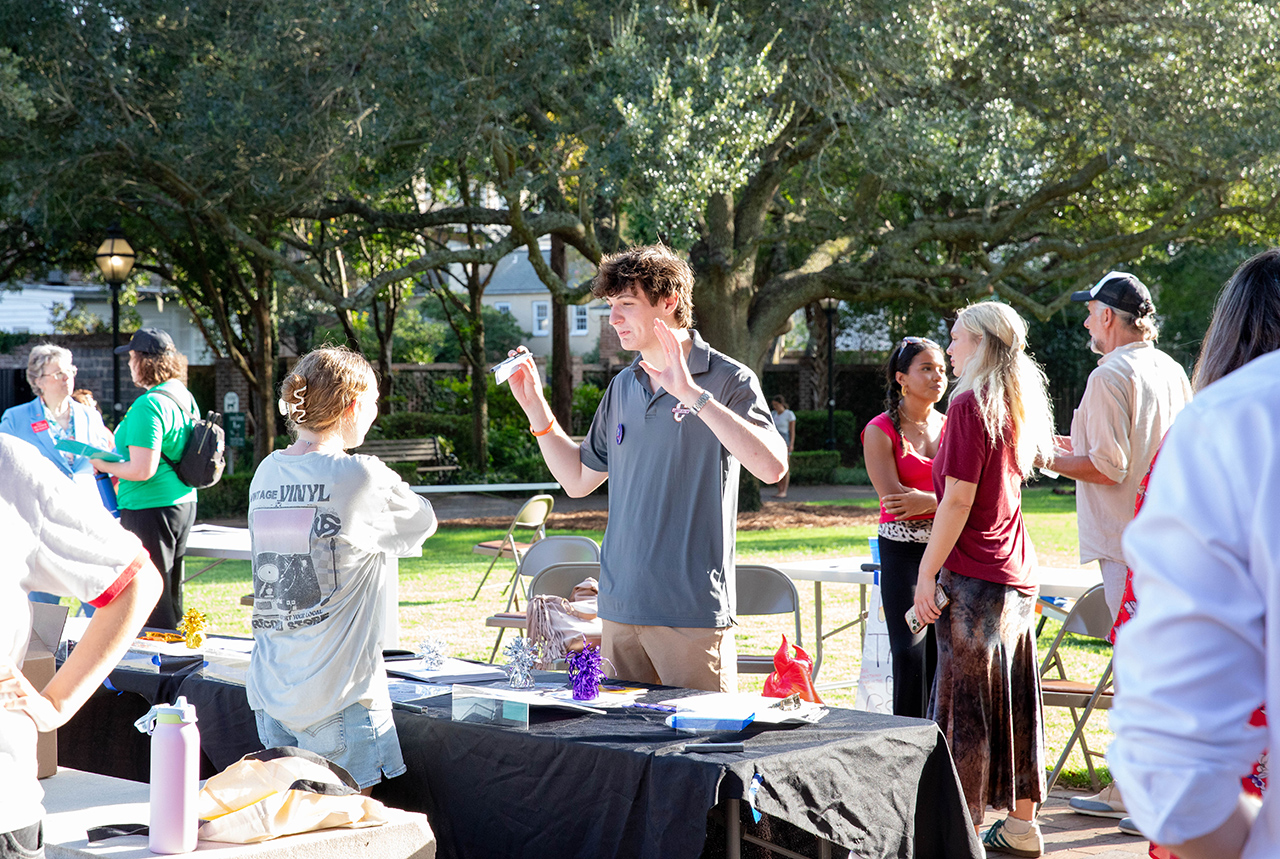 A young person gestures while speaking at an outdoor event, surrounded by attendees and tables set up under large trees.