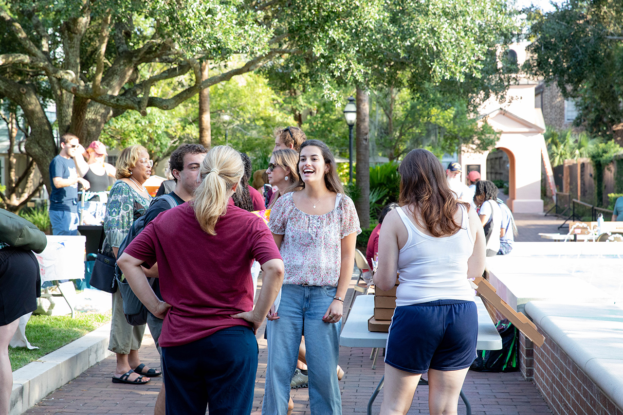 A group of people socialize outdoors under trees, with one person laughing joyfully while others engage in conversation.
