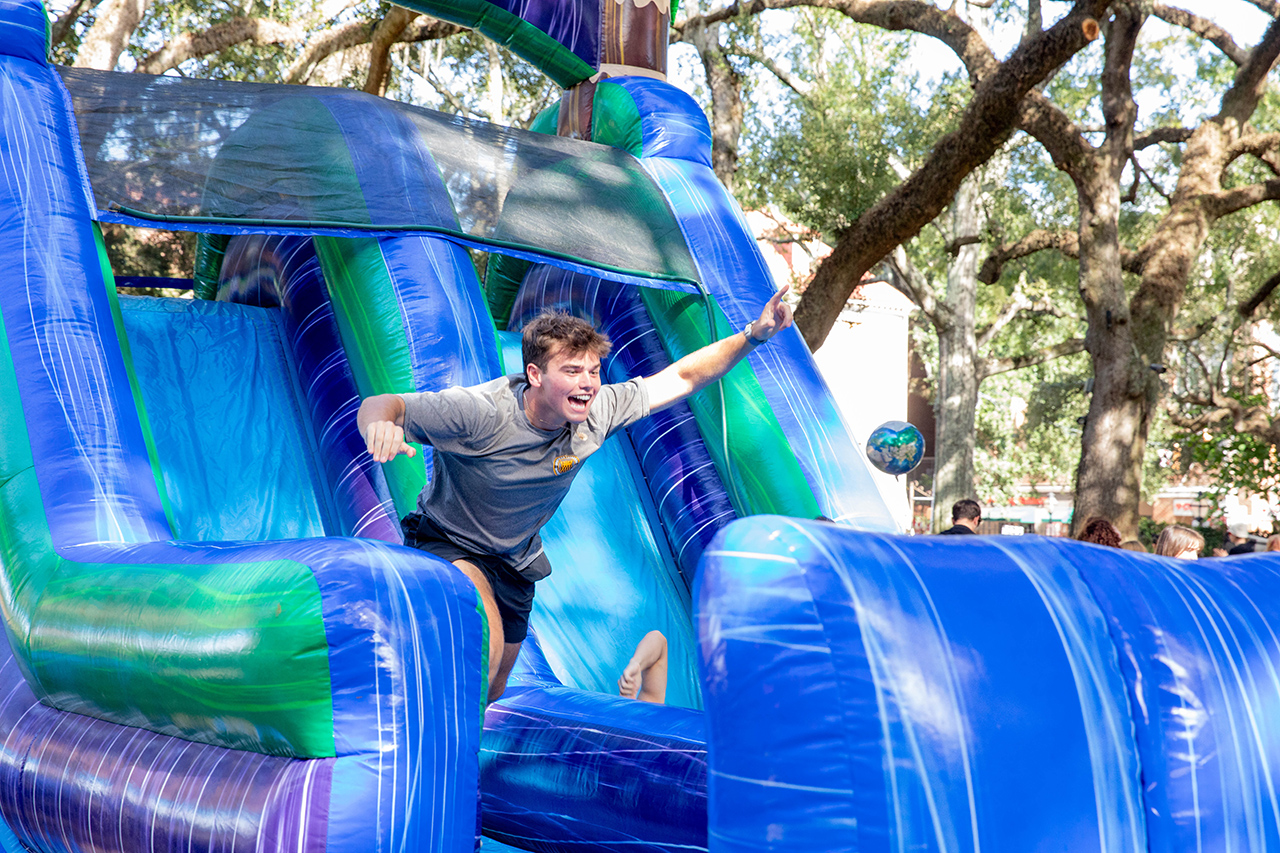 A person slides down a colorful inflatable slide outdoors, arms outstretched and smiling.