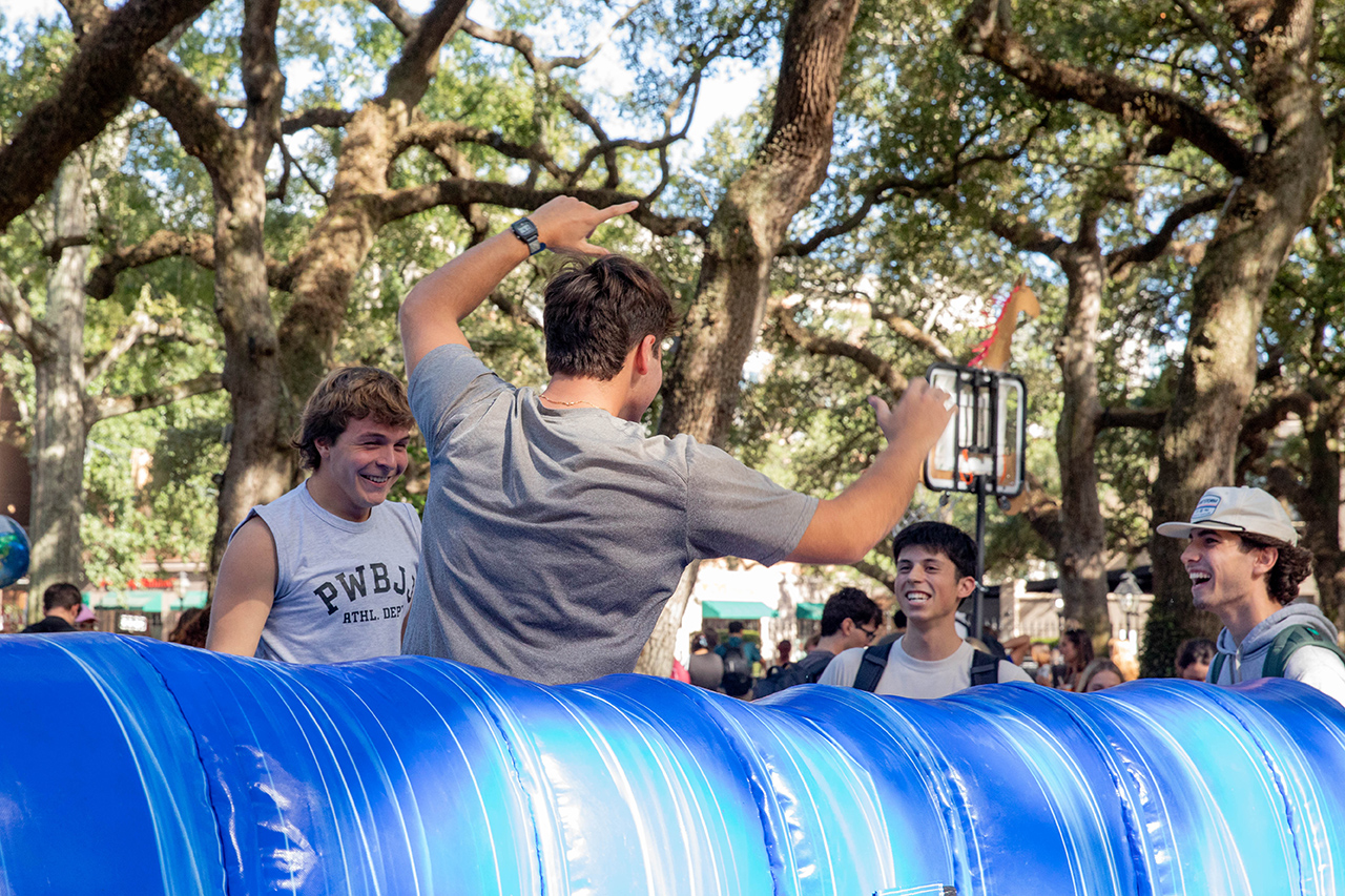 Several people laugh and gesture around a large blue inflatable structure under leafy trees.
