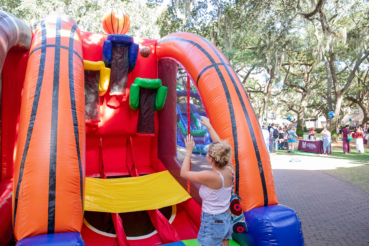 A person interacts with a colorful inflatable basketball game in an outdoor setting surrounded by trees and other attendees.