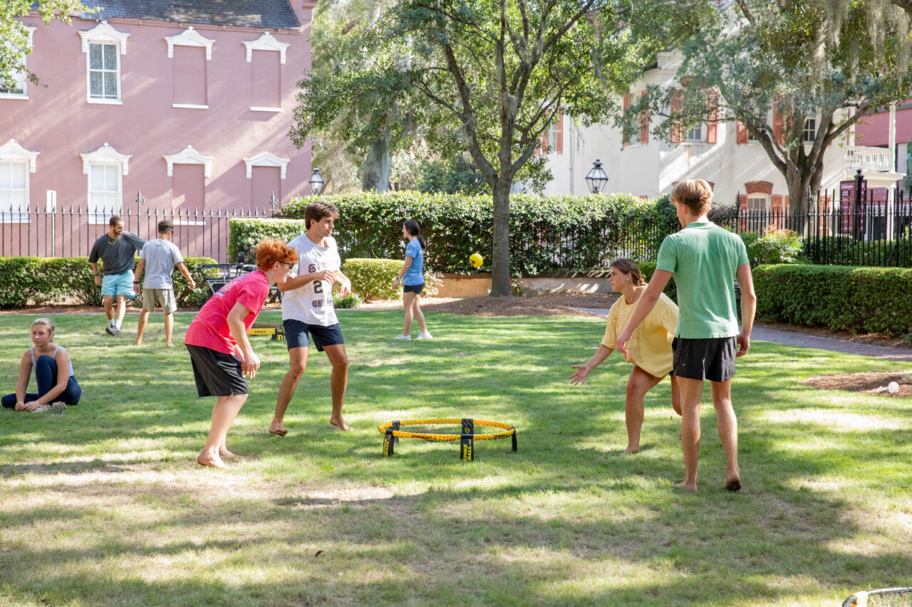 A group of young people play a game of ring toss on a sunny lawn in front of historic buildings.
