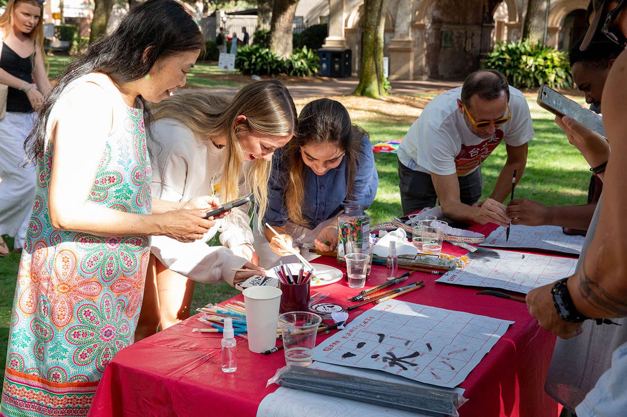 A group of people gather around a table covered with a red cloth, engaging in an activity that involves writing or drawing with brushes and ink on paper.