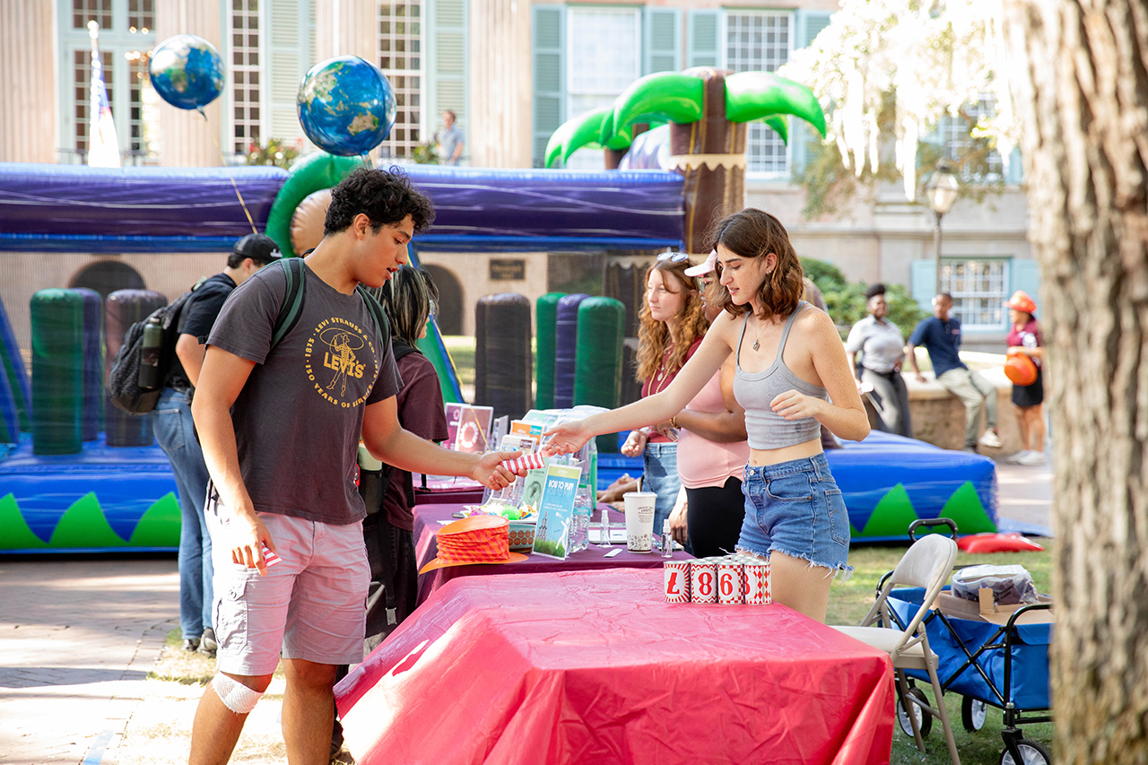 Two individuals interact at a table covered with a red tablecloth, surrounded by inflatable play structures and balloons in an outdoor setting.