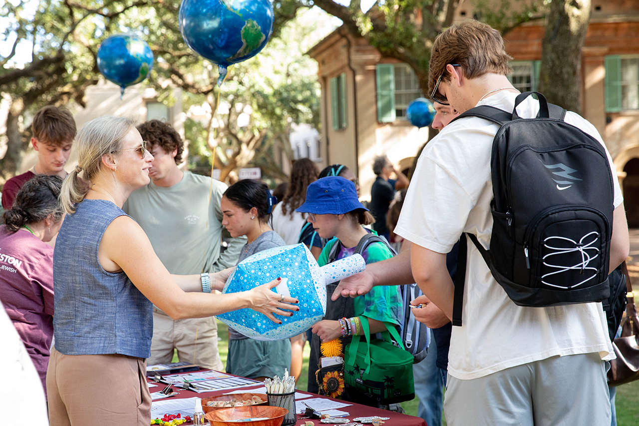 A group of people gather outdoors under trees, interacting around a table with a large blue and white patterned object, while balloons shaped like globes float above.
