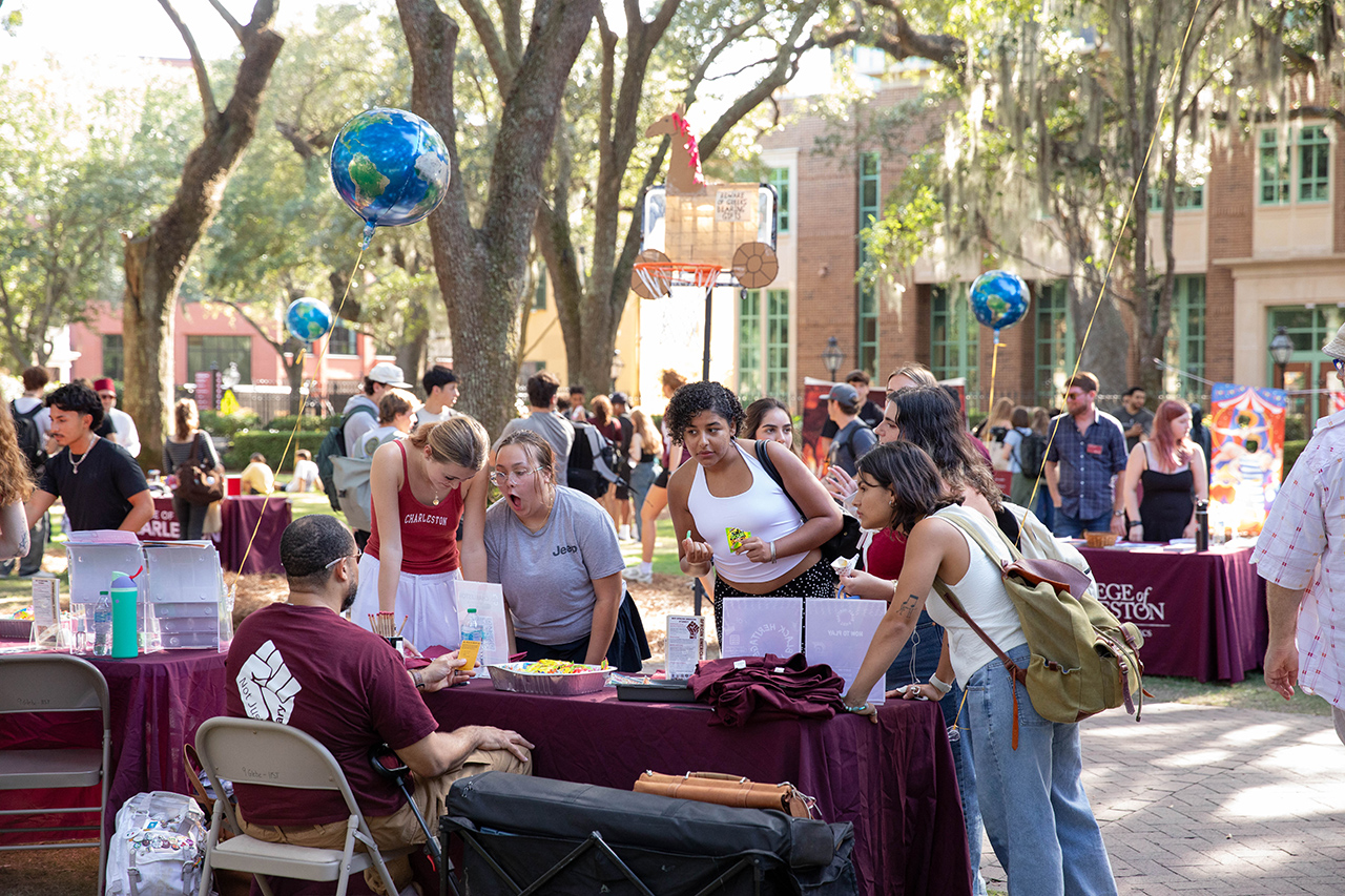 Students gather around a table at an outdoor event, engaging with a representative under trees and colorful balloons.