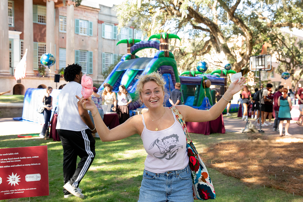 A smiling person holds up cotton candy and a ticket at an outdoor event with inflatable slides and people in a grassy area.