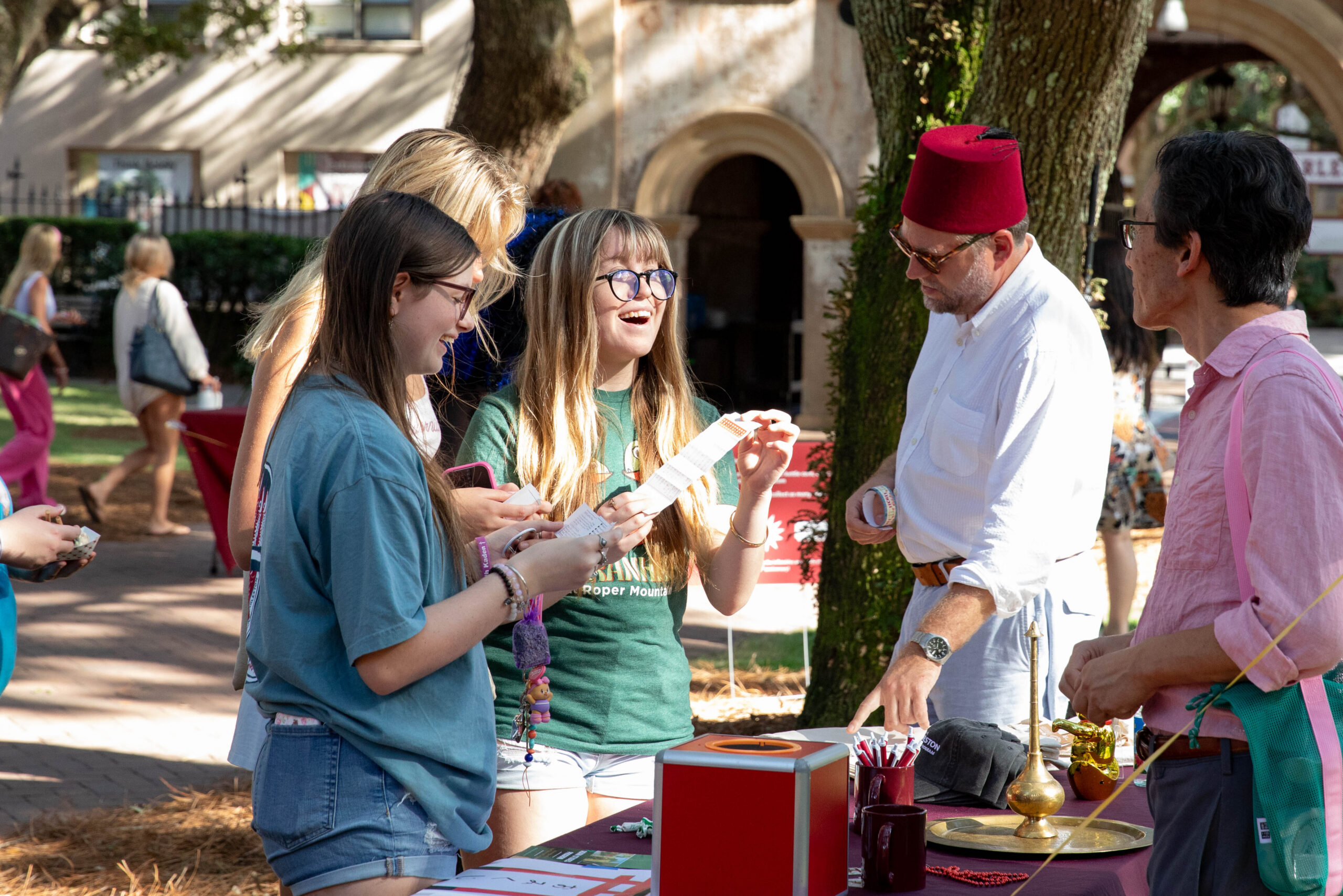 A group gathers around a table outdoors under trees, where one person in a red fez interacts with others holding papers, while a table displays items including a red box and a brass lamp.