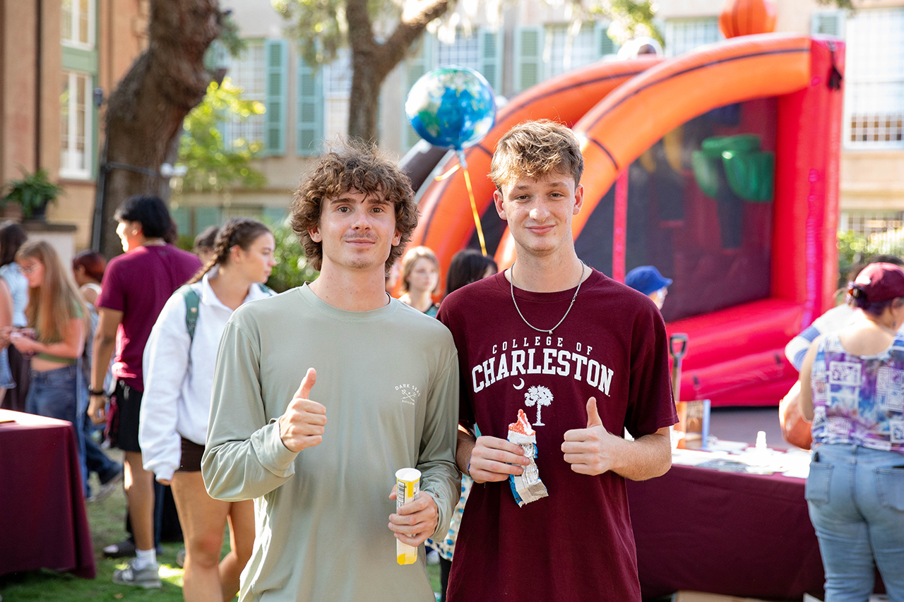 Two young people smile and give thumbs-up gestures at an outdoor event with a bounce house and balloons in the background.