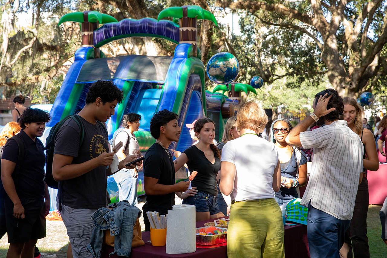 A group of people socialize outdoors near a large inflatable water slide under trees.
