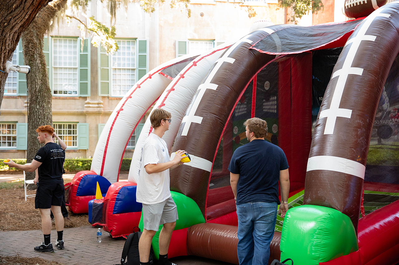 Three individuals interact near a large inflatable football-themed game structure outdoors.