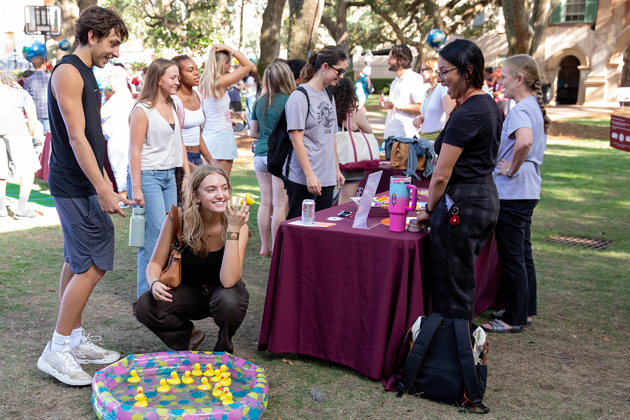 A group of young people gather outdoors around a table with a purple tablecloth, where one person is crouching beside a colorful inflatable pool filled with yellow rubber ducks.