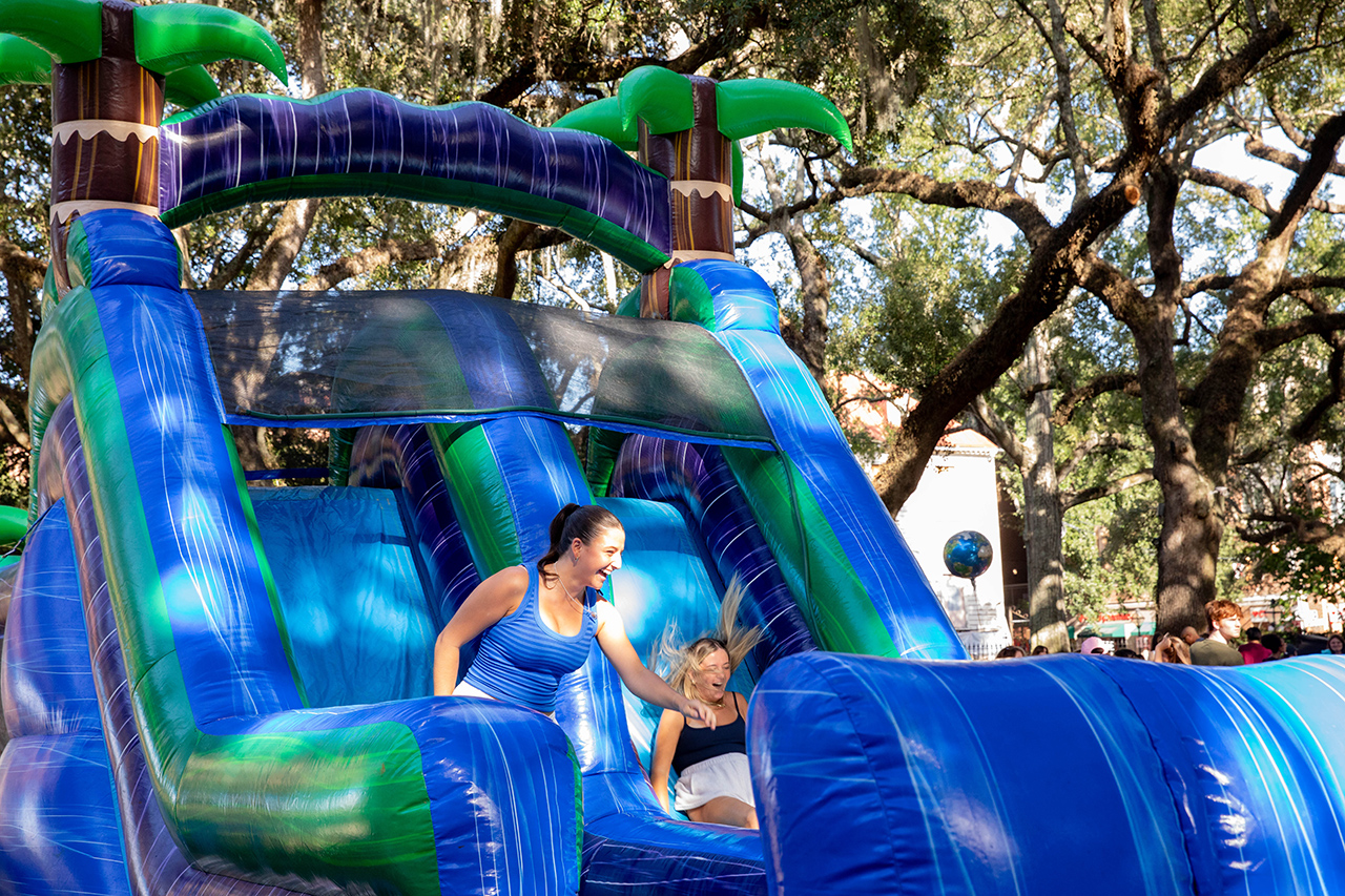 Two people enjoy sliding down a colorful inflatable water slide under a canopy of trees.