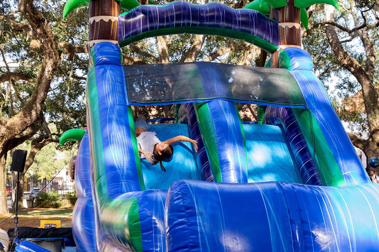 A person slides down a large blue and green inflatable water slide outdoors, surrounded by trees.