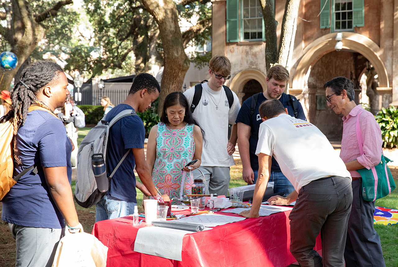 A group of people gathers around a red-clothed table outdoors, engaging with materials and each other under the shade of large trees near a historic building.