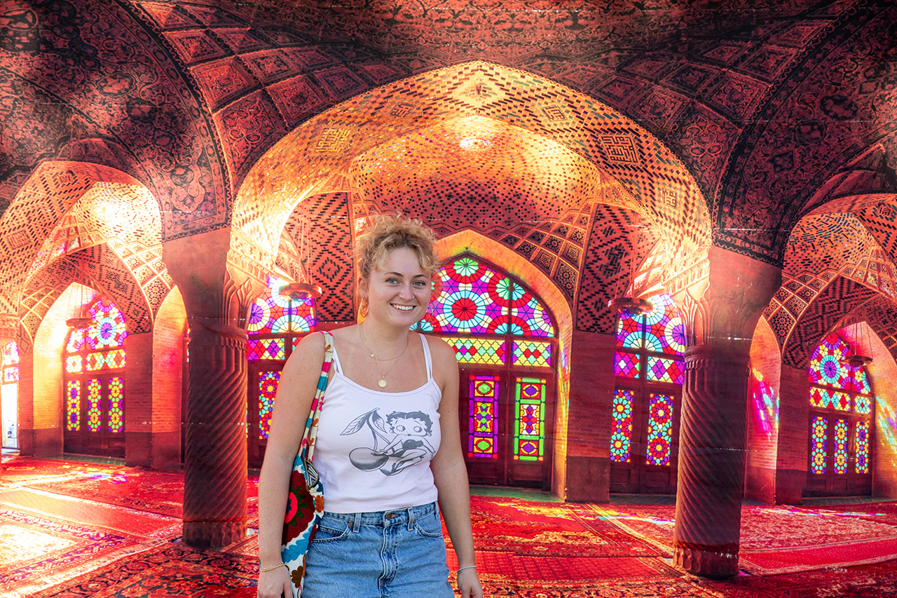 A person smiles in a vibrant, ornately decorated hall with intricate arches, stained glass windows, and richly patterned carpets.