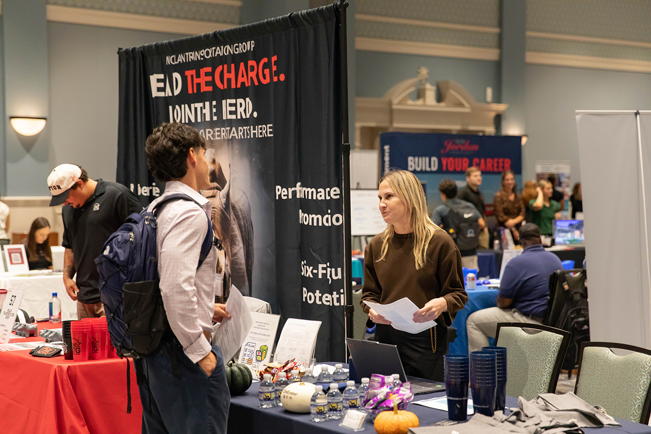 A person with a backpack engages in conversation with a recruiter at a job fair booth, surrounded by other attendees and career-related displays.