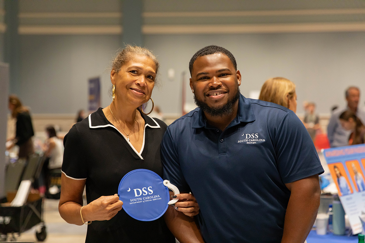 Two individuals smile for the camera at an event, one holding a blue circular sign with “DSS South Carolina” branding.