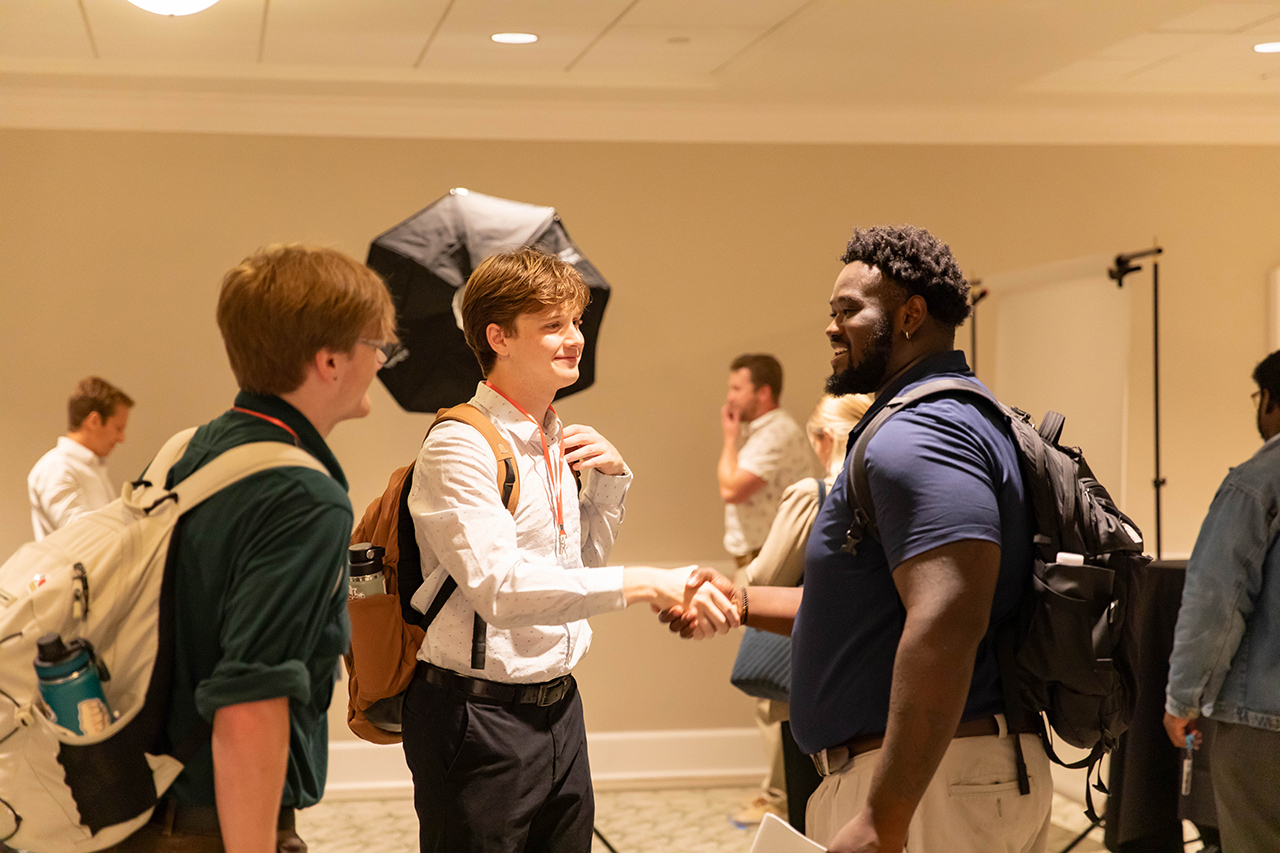 Two individuals shake hands in a warmly lit indoor space, surrounded by others and photography equipment.