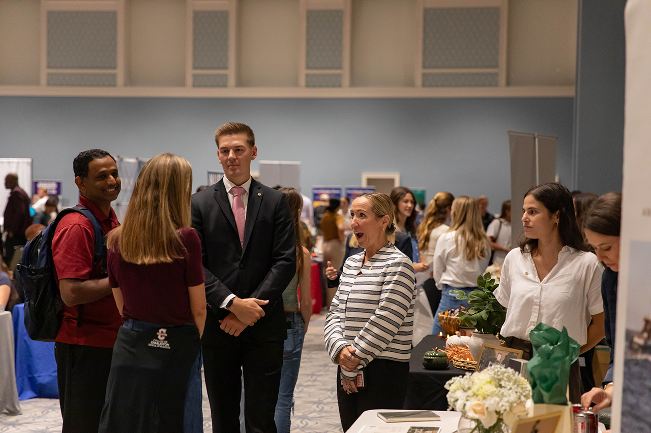 A group of people engage in conversation at an indoor event, with some standing near a table displaying floral arrangements and informational materials.