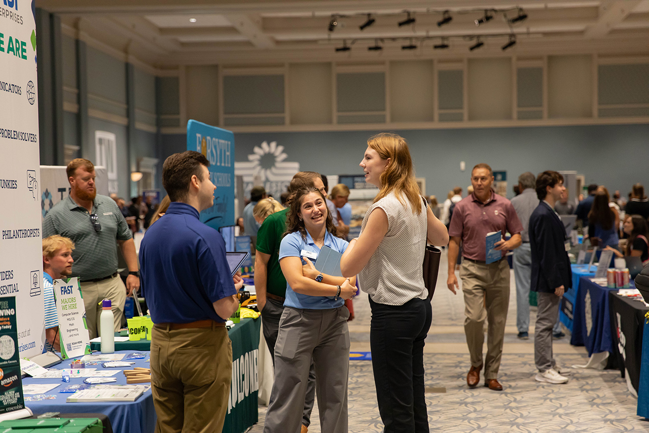 Attendees engage in conversation at a career fair with booths displaying company information and promotional materials.
