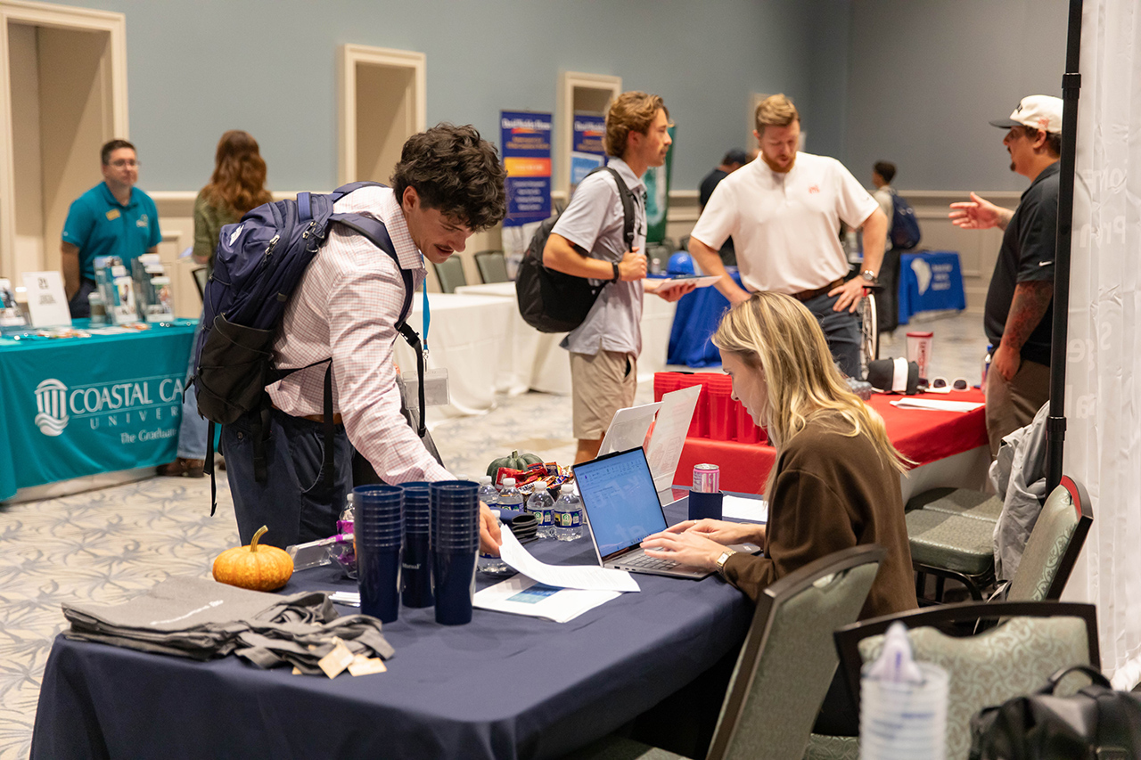 A person with a backpack leans over a table at a college fair booth, interacting with a seated representative who is using a laptop.