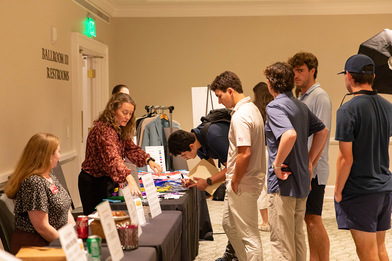 A group of people gather around a table with merchandise and informational materials at an event in a ballroom.