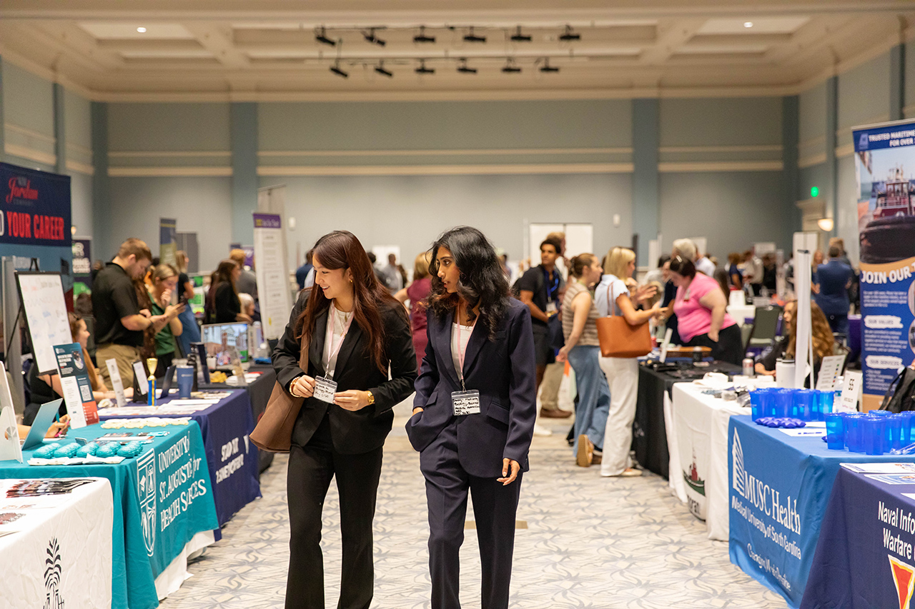 Two individuals in professional attire walk through a bustling career fair, engaging with booths and networking among attendees.