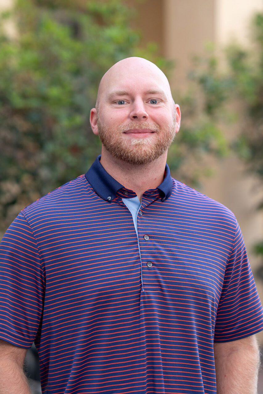 A bald person with a beard smiles while wearing a striped polo shirt outdoors.