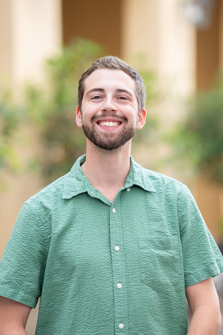 A smiling person in a green button-up shirt stands outdoors with blurred greenery and buildings in the background.
