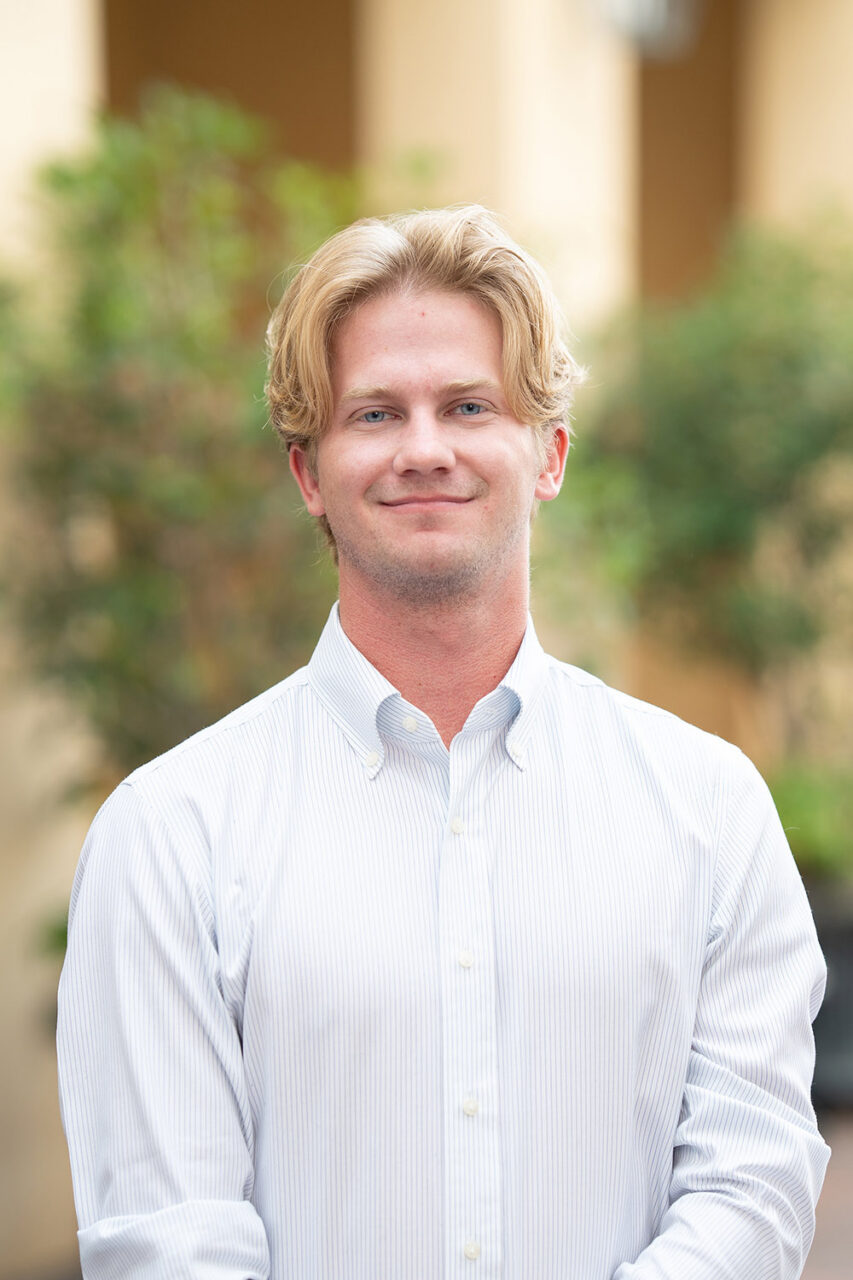 A person with light-colored hair stands outdoors in a white button-down shirt, smiling gently at the camera against a softly blurred background of greenery and building columns.