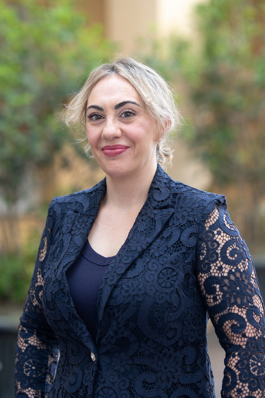 A person with blonde hair smiles while wearing a dark blue lace blazer outdoors, with greenery and benches in the background.