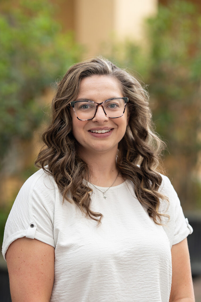 A person with curly hair and glasses smiles while wearing a white shirt outdoors.