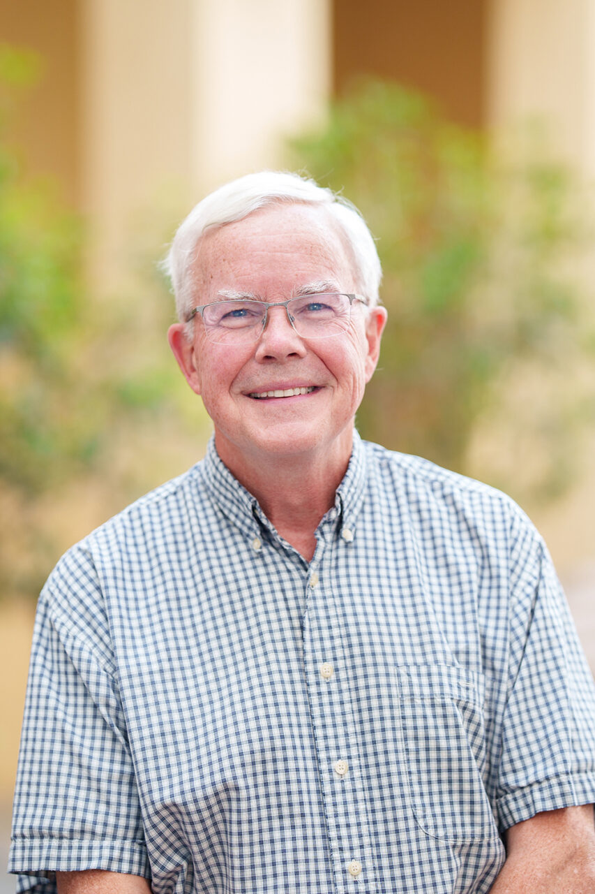 An older person with white hair and glasses smiles while wearing a short-sleeved checkered shirt, standing outdoors in front of blurred greenery and columns.
