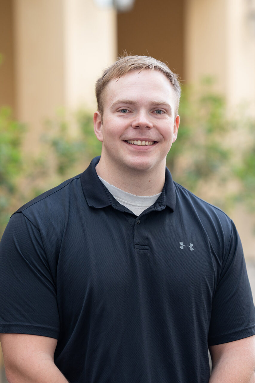 A smiling person in a black polo shirt stands outdoors in front of a building with columns and greenery.