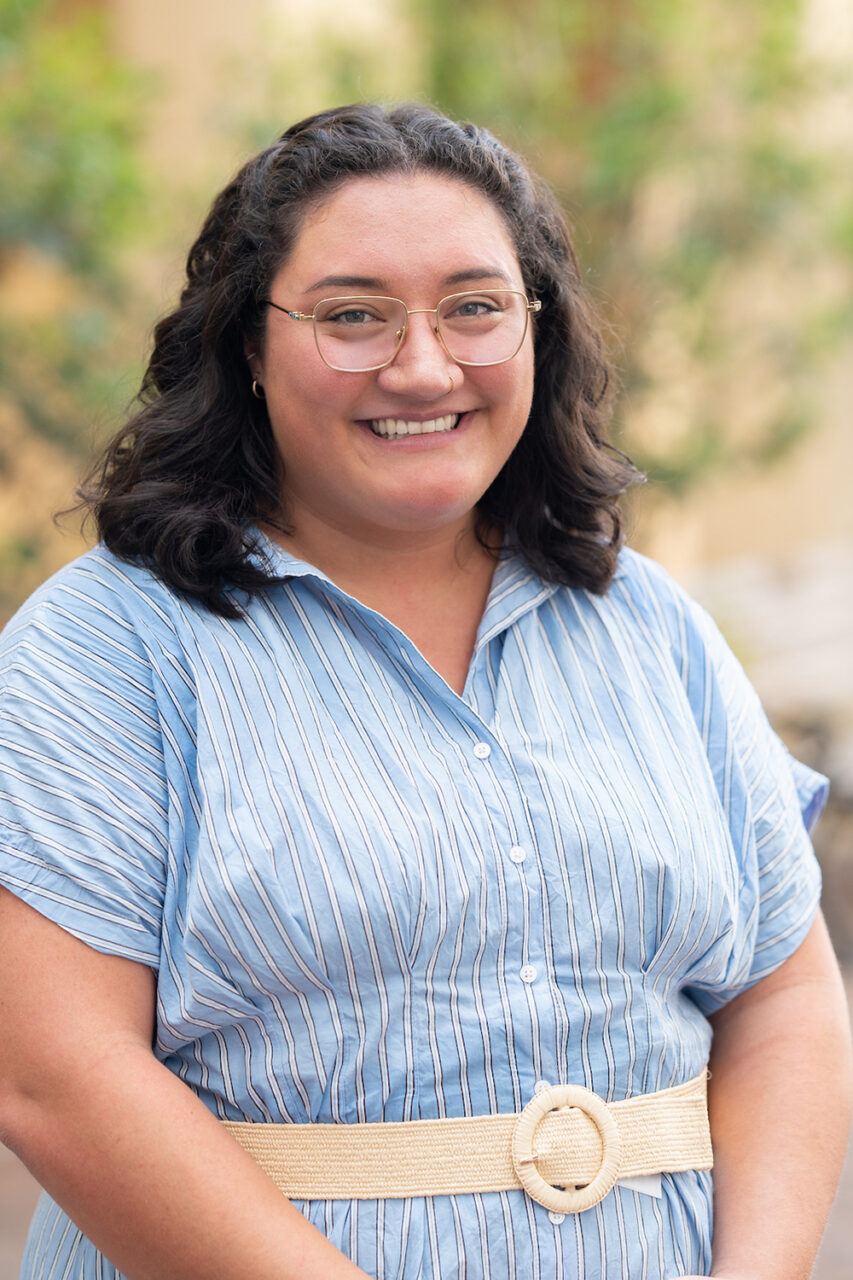 A smiling person with dark curly hair and glasses stands outdoors in a blue striped dress, wearing a woven belt, with greenery and blurred background elements.