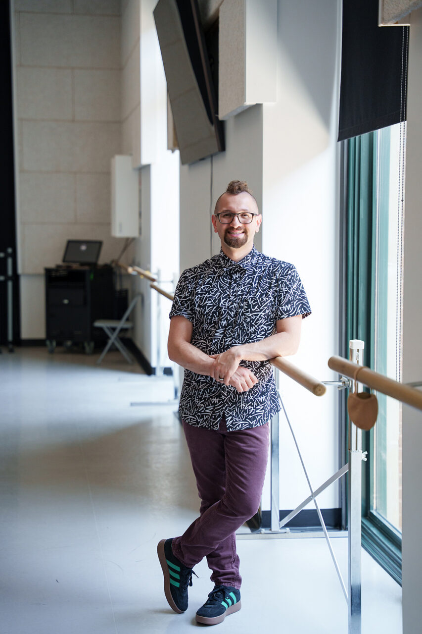 A person with glasses and a mohawk stands smiling, leaning against a ballet barre in a bright, modern studio.