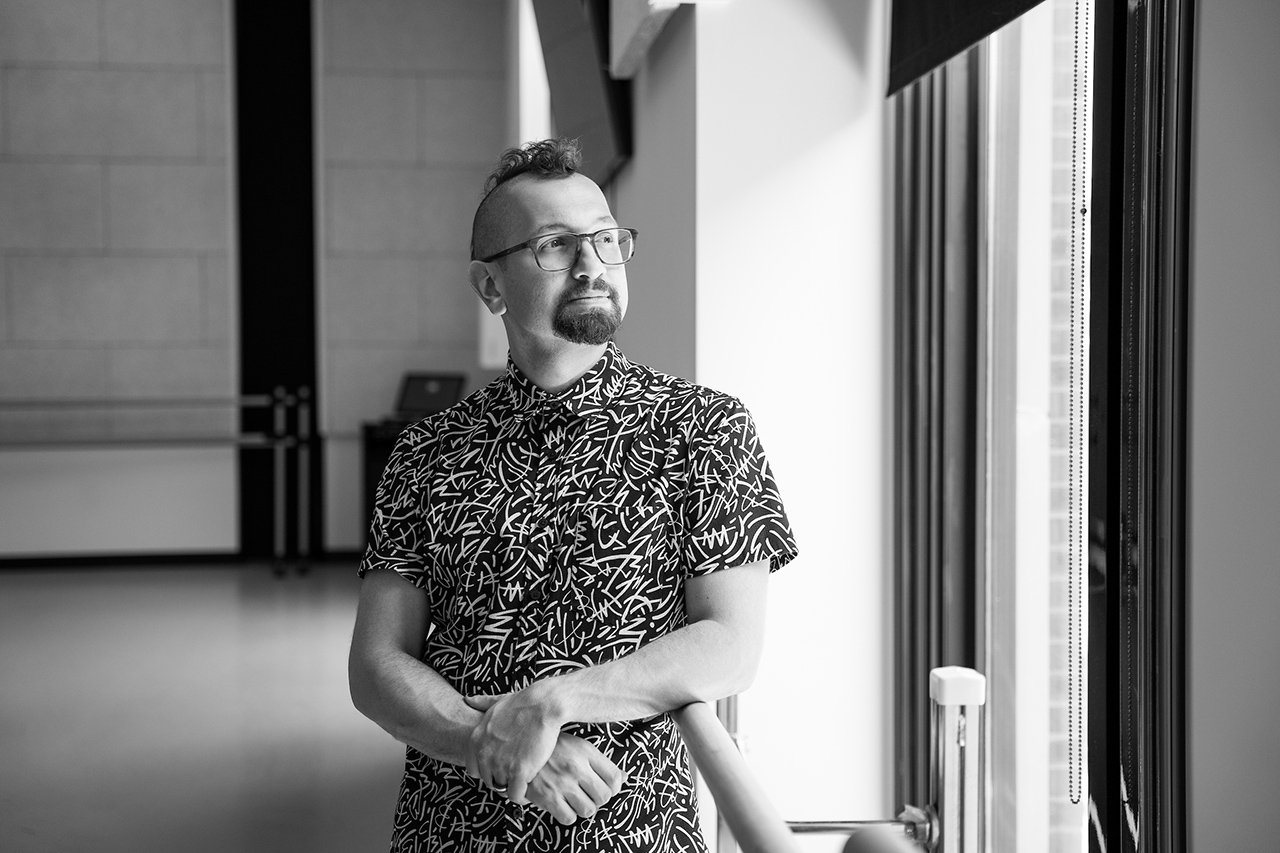 A person with glasses and a patterned shirt stands near a window, gazing outward with hands clasped.