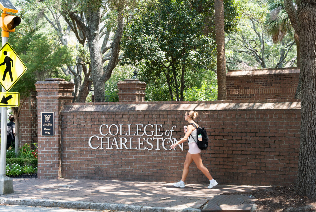 A person walks past a brick wall bearing the sign "College of Charleston" on a sunny day, with trees and traffic signs visible nearby.